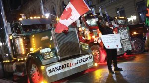 Convoy trucks on an Ottawa street at night with a Canadian flag and a "Mandate Freedom" banner, beside a protester holding a sign